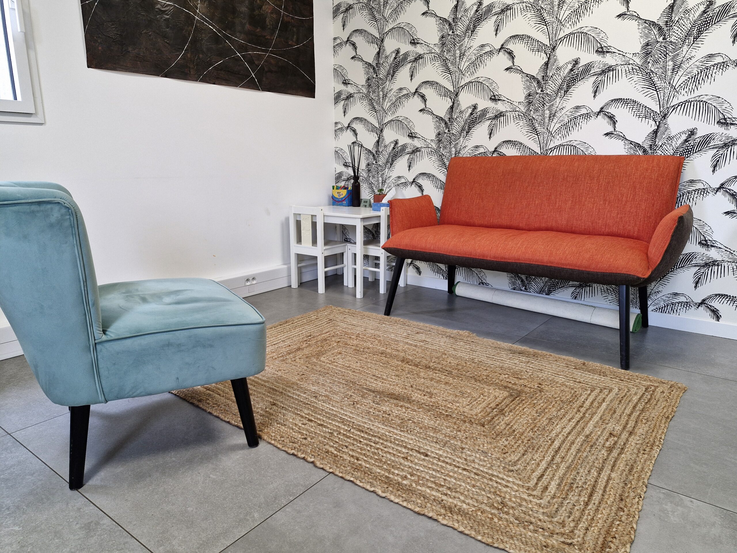 Cabinet de psychologue avec un fauteuil bleu, un canapé orange, un tapis en fibres naturelles et un coin pour enfants avec une petite table et deux chaises blanches, sur fond de papier peint à motifs de palmiers