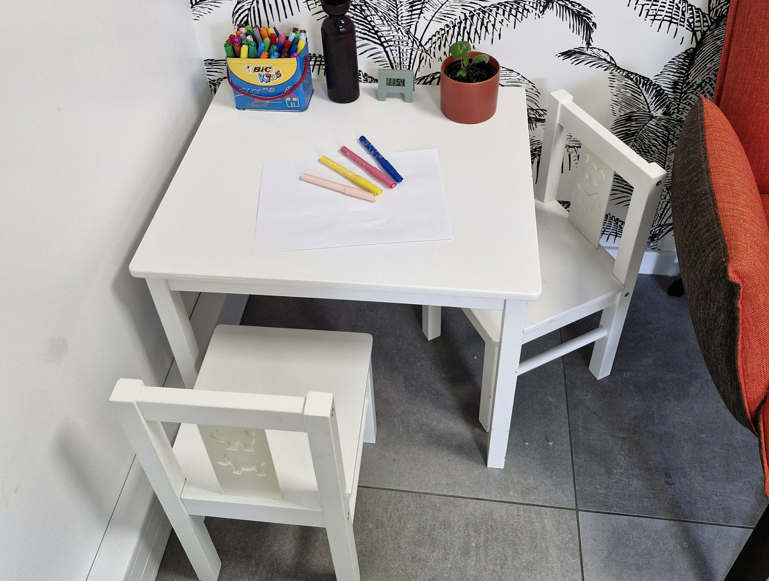 Petite table pour enfants avec deux chaises blanches. Sur la table se trouvent une feuille, des feutres colorés, une boîte de crayons et une petite plante en pot.