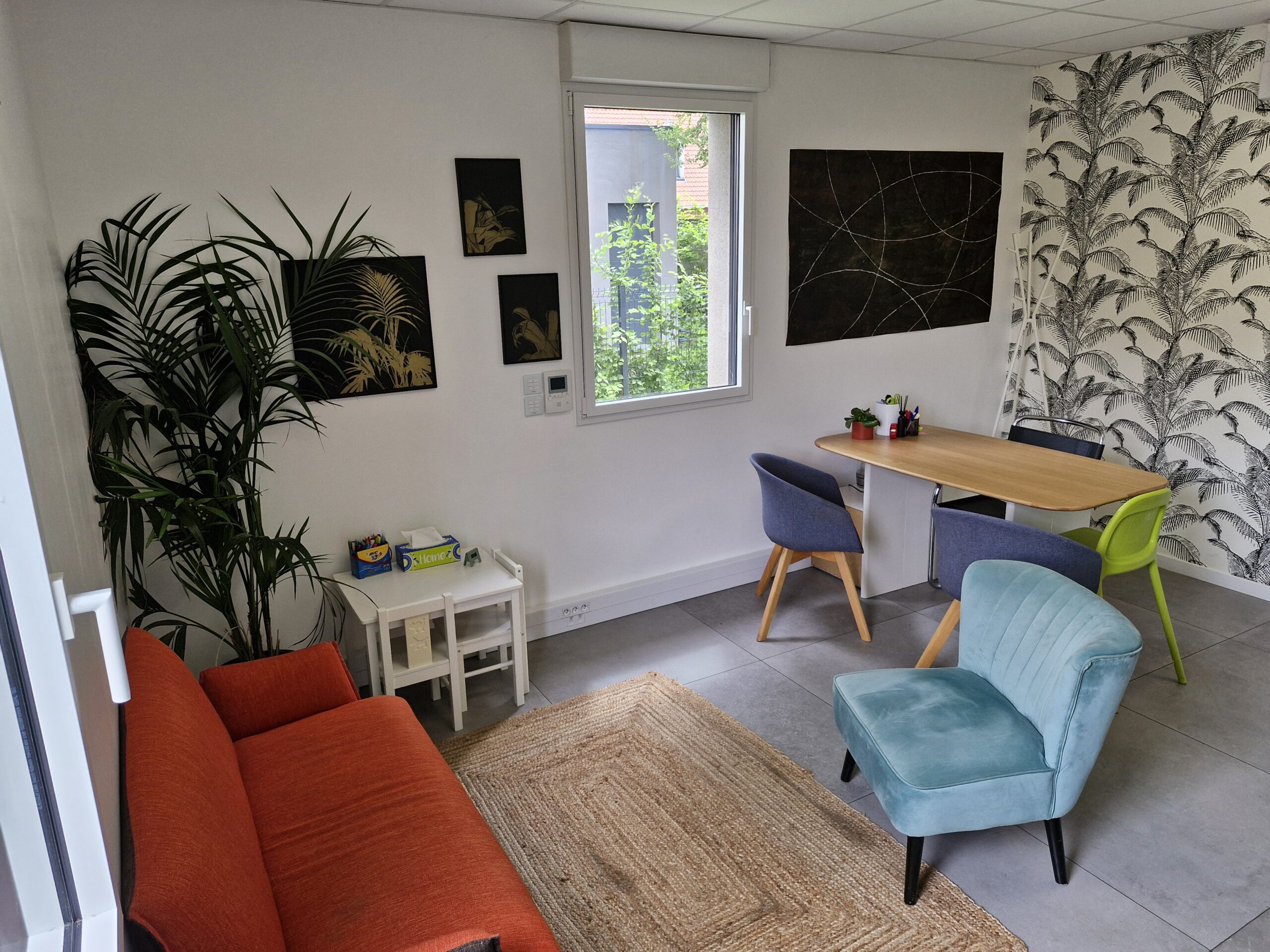 Bureau de consultation lumineux avec une décoration moderne. Un canapé orange, un fauteuil vert sauge, un tapis en fibres naturelles, une table pour enfants avec deux petites chaises et un espace bureau sont disposé dans la pièce. Dans le coin, une grande plante verte apporte une touche végétale.