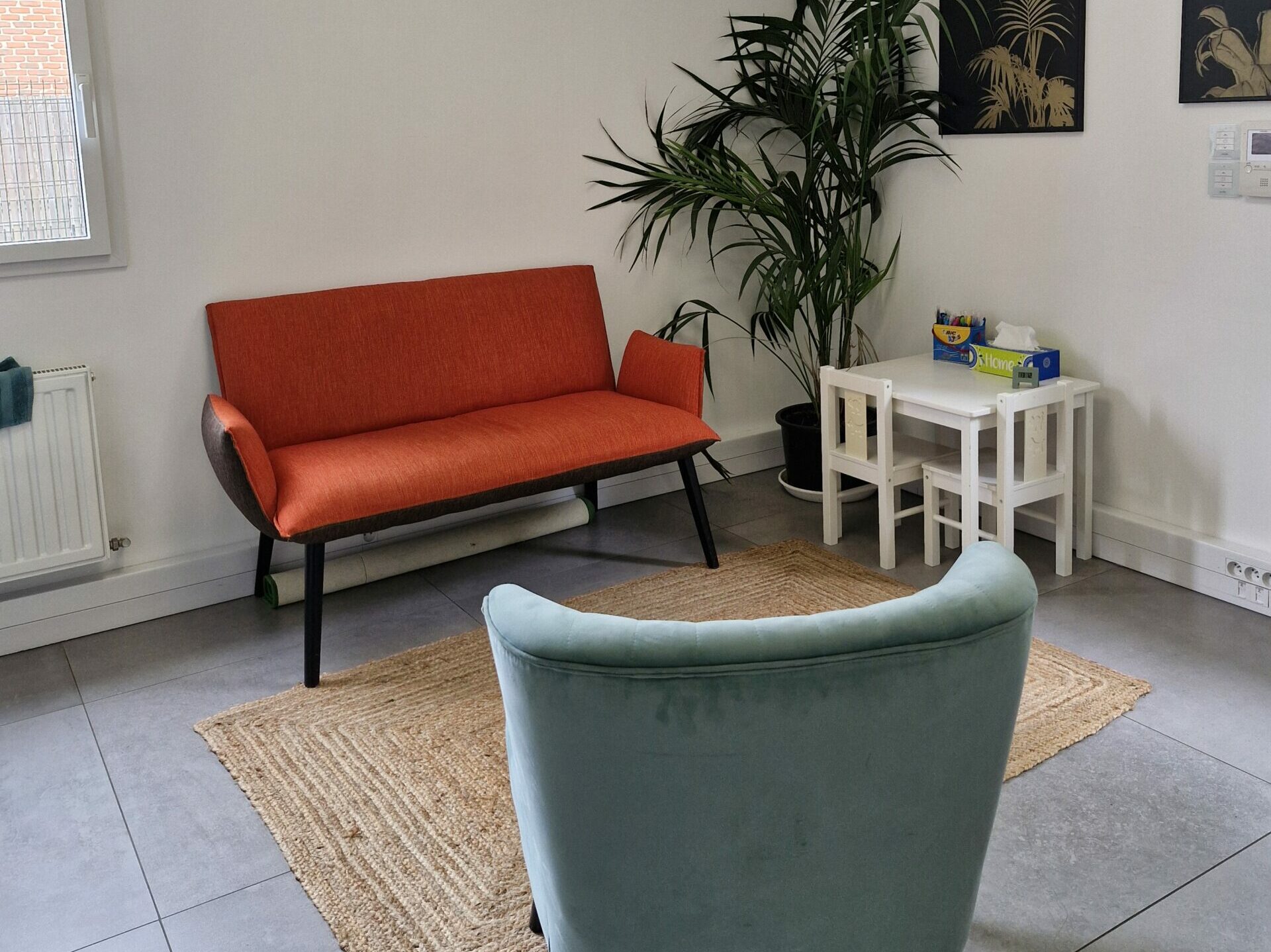 Bureau de consultation au décor chaleureux et minimaliste : un canapé orange, un fauteuil vert sauge, un tapis en fibres naturelles, une table pour enfants avec deux petites chaises. Dans le coin, une grande plante verte apporte une touche végétale.